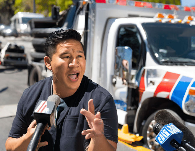 A news reporter broadcasts live from a San Jose street, highlighting a towing truck assisting drivers.